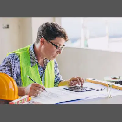 Construction professional in safety vest calculating costs on a tablet while reviewing building plans.