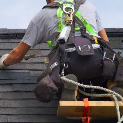 Professional roofer in safety harness installing new shingles on a residential roof, equipped with various roofing tools and equipment.