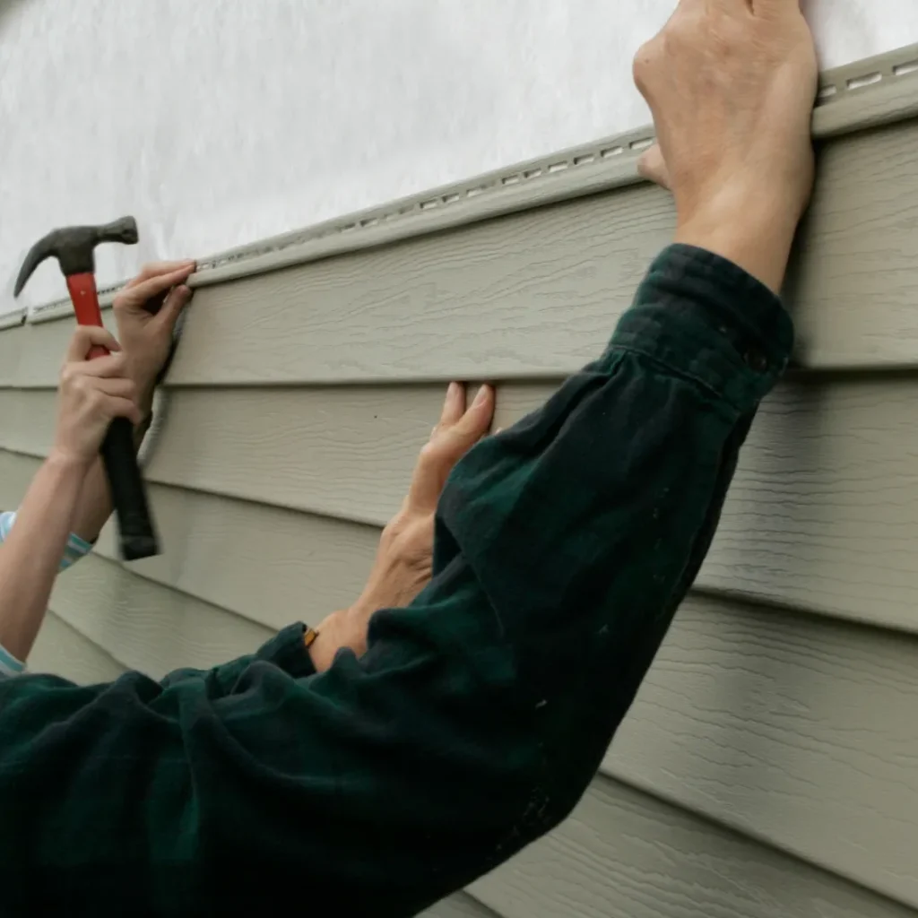 Two workers installing vinyl siding on the side of a home using a hammer.